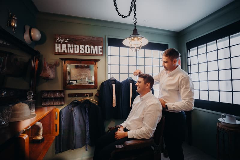 The groom sits in a chair while a man styles his hair in a room at Preston Peak Winery Functions Centre, with suits and ties hanging on the wall and a sign that reads 'keep it HANDSOME'.