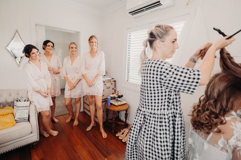 The bride Chelsea is having her hair styled by a hairdresser while four bridesmaids in matching robes watch in a room at Preston Peak Winery Functions Centre.