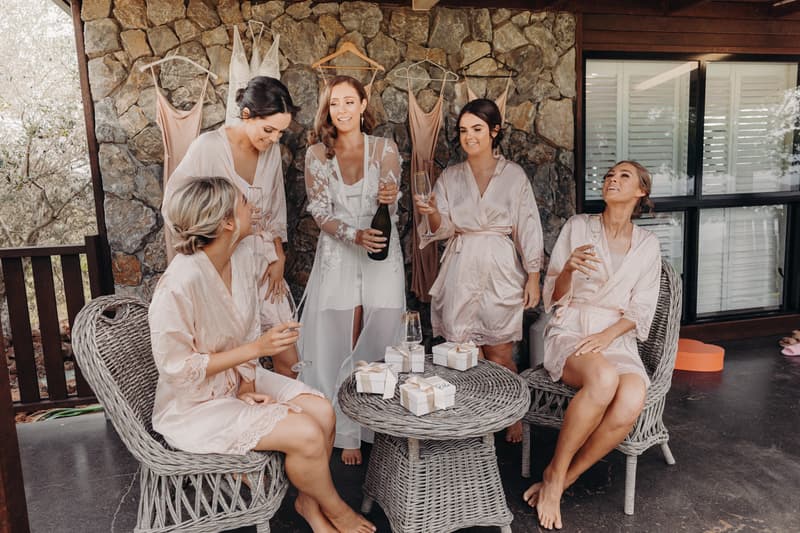 The bride Chelsea and four bridesmaids in matching robes gather around a wicker table on a covered patio at Preston Peak Winery Functions Centre. Chelsea is holding a bottle of champagne while the bridesmaids hold champagne flutes. Wedding dresses and bridesmaid dresses hang on the stone wall behind them.