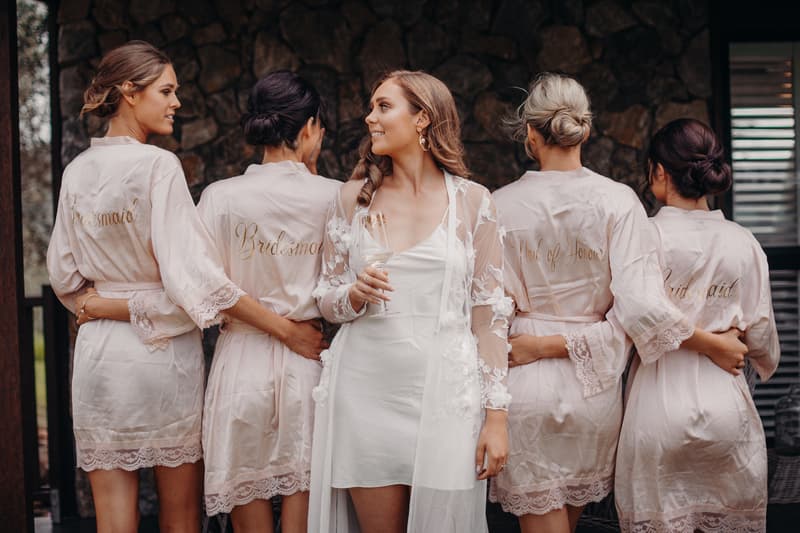 The bride Chelsea in a white dress and sheer floral robe holds a glass while standing with four bridesmaids and a maid of honor wearing matching light pink robes with gold lettering at Preston Peak Winery Functions Centre.