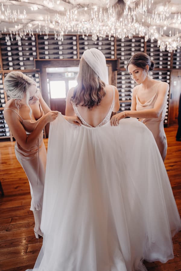 The bride Chelsea stands with her back to the camera wearing a wedding gown and veil while two bridesmaids adjust the train of her dress inside Preston Peak Winery Functions Centre.