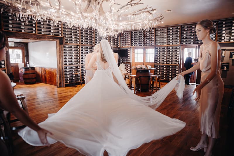 The bride at Preston Peak Winery Functions Centre is assisted by bridesmaids as they hold and arrange her flowing wedding dress train indoors in a room lined with wine racks.