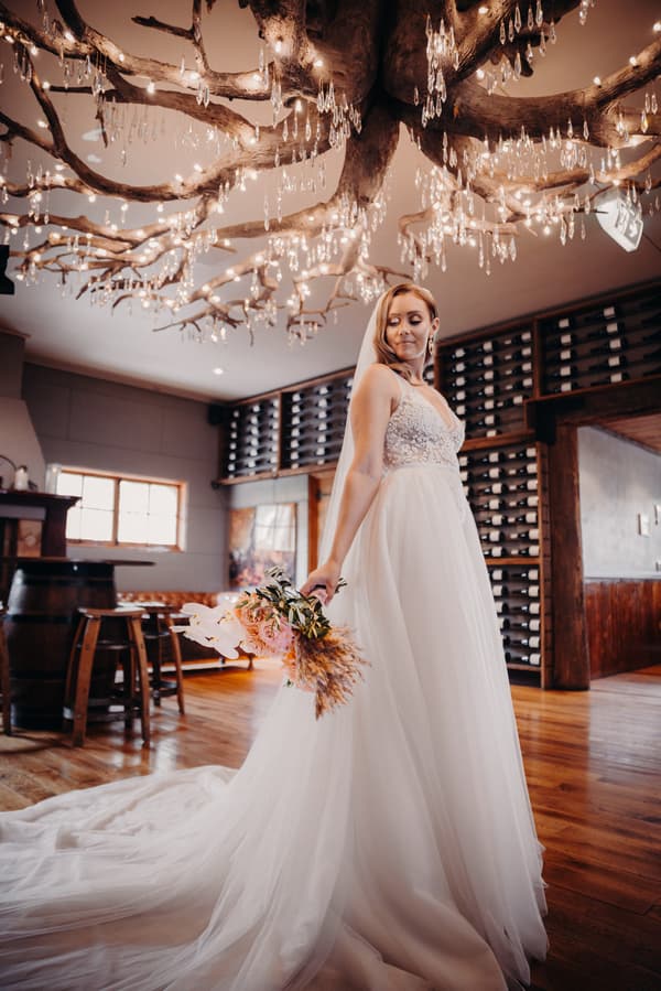 Chelsea, the bride, poses in her wedding gown holding a bouquet inside Preston Peak Winery Functions Centre, with a decorative chandelier and wine racks visible in the background.