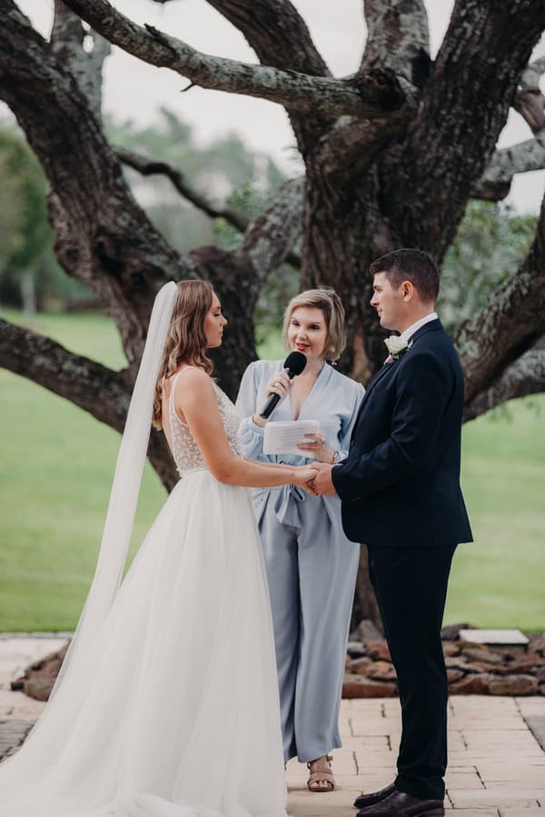 Bride Chelsea and groom Brent hold hands facing each other while an officiant in a light blue outfit speaks into a microphone during their ceremony at Preston Peak Winery Functions Centre outdoors in front of a large tree.