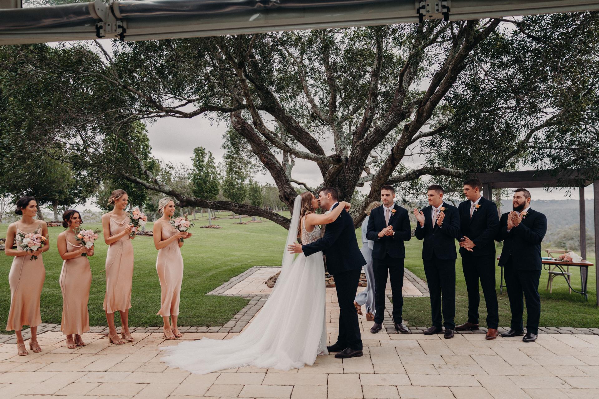 Bride Chelsea and groom Brent kiss at the ceremony stage at Preston Peak Winery Functions Centre, surrounded by bridesmaids in peach dresses holding bouquets and groomsmen in dark suits applauding under a large tree.
