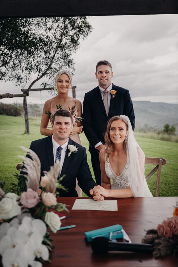 Chelsea the bride and Brent the groom sit at a table holding hands with a signed document in front of them at Preston Peak Winery Functions Centre. Behind them stand a bridesmaid and a groomsman outdoors with greenery and hills in the background.