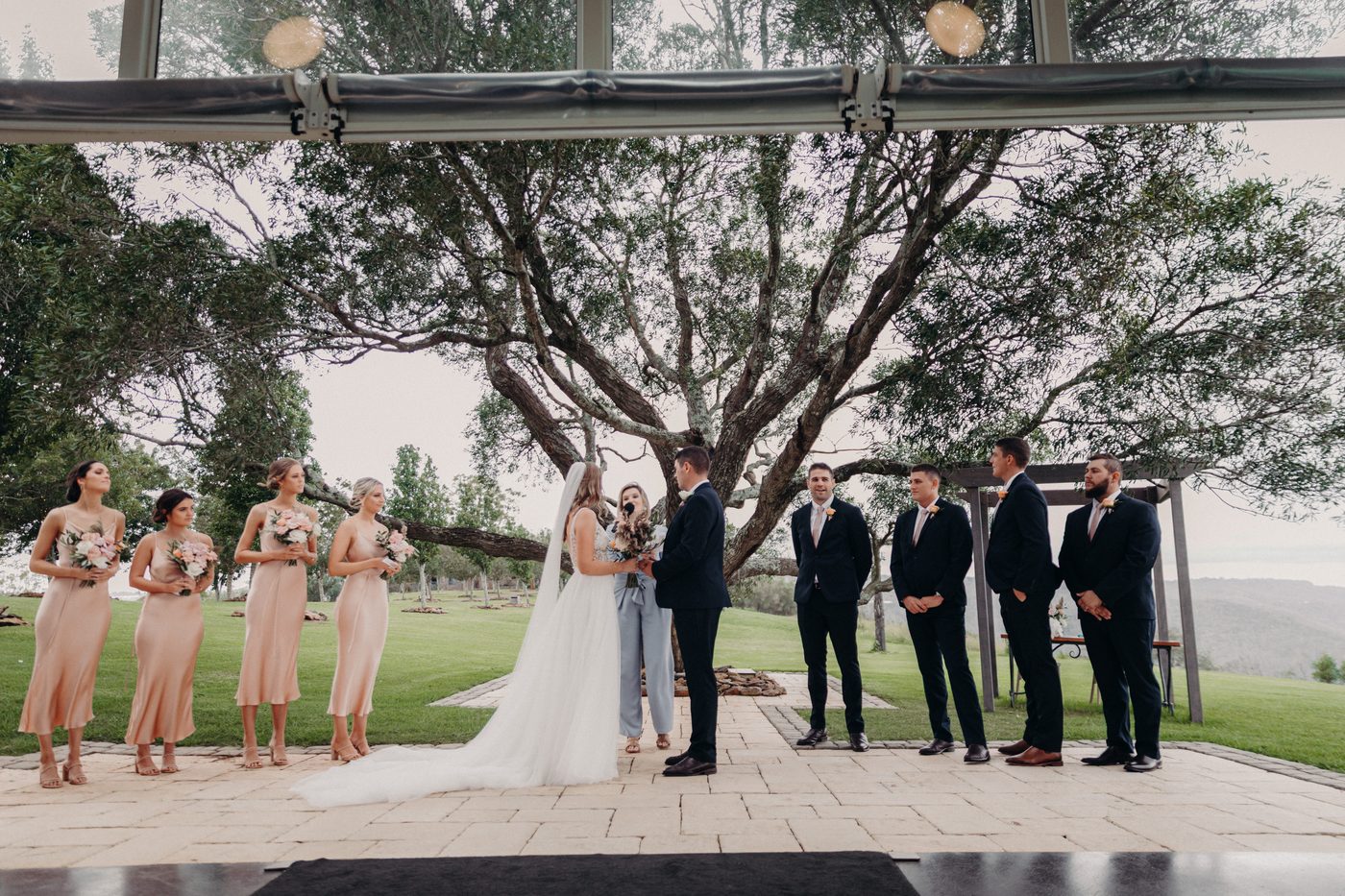Chelsea and Brent stand at the ceremony stage, holding hands and facing each other while the celebrant speaks. Four bridesmaids in matching light peach dresses hold bouquets on the left, and five groomsmen in dark suits stand on the right under a large tree outdoors. Engagement celebration by She Said Yes.
