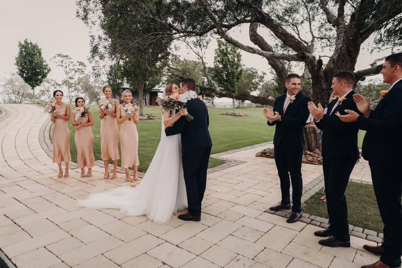 Bride Chelsea and groom Brent kiss at the Preston Peak Winery Functions Centre ceremony stage while bridesmaids in matching dresses hold bouquets and groomsmen in suits applaud.
