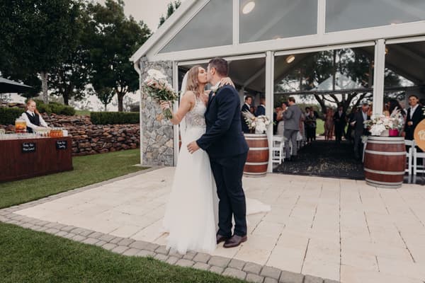 Bride Chelsea and groom Brent kiss outside the Preston Peak Winery Functions Centre after their ceremony, with guests visible inside the venue and a server preparing drinks at a nearby table.