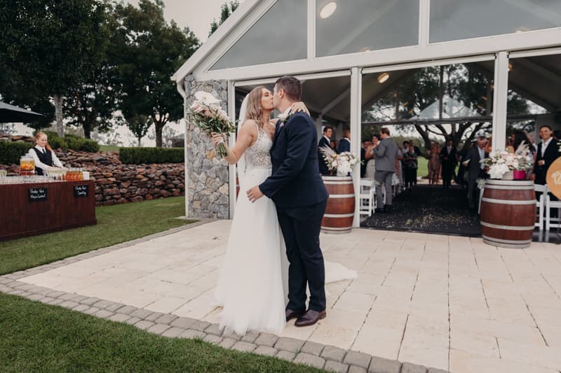 Bride Chelsea and groom Brent kiss outside the Preston Peak Winery Functions Centre after their ceremony, with guests visible inside the venue and a server preparing drinks at a nearby table.