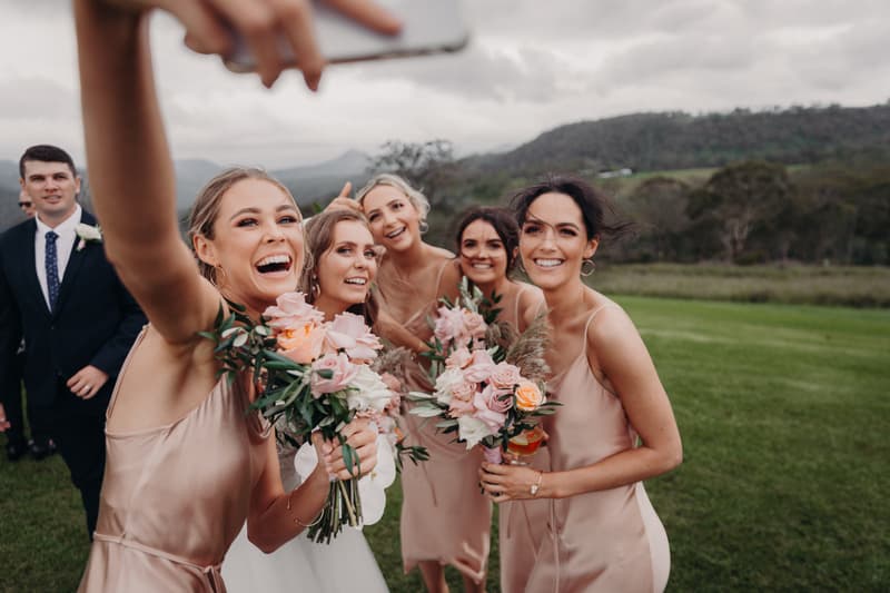 The bride Chelsea and four bridesmaids in matching blush dresses hold bouquets and pose for a selfie outdoors at Preston Peak Winery Functions Centre, with a man in a suit visible in the background.