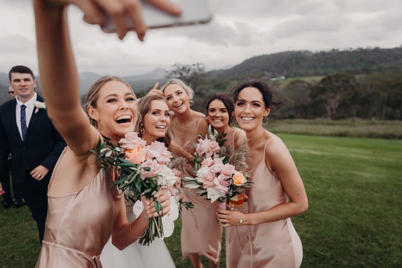 The bride Chelsea and four bridesmaids in matching blush dresses hold bouquets and pose for a selfie outdoors, with a man in a suit visible in the background. Wedding photography pricing estimate by She Said Yes.