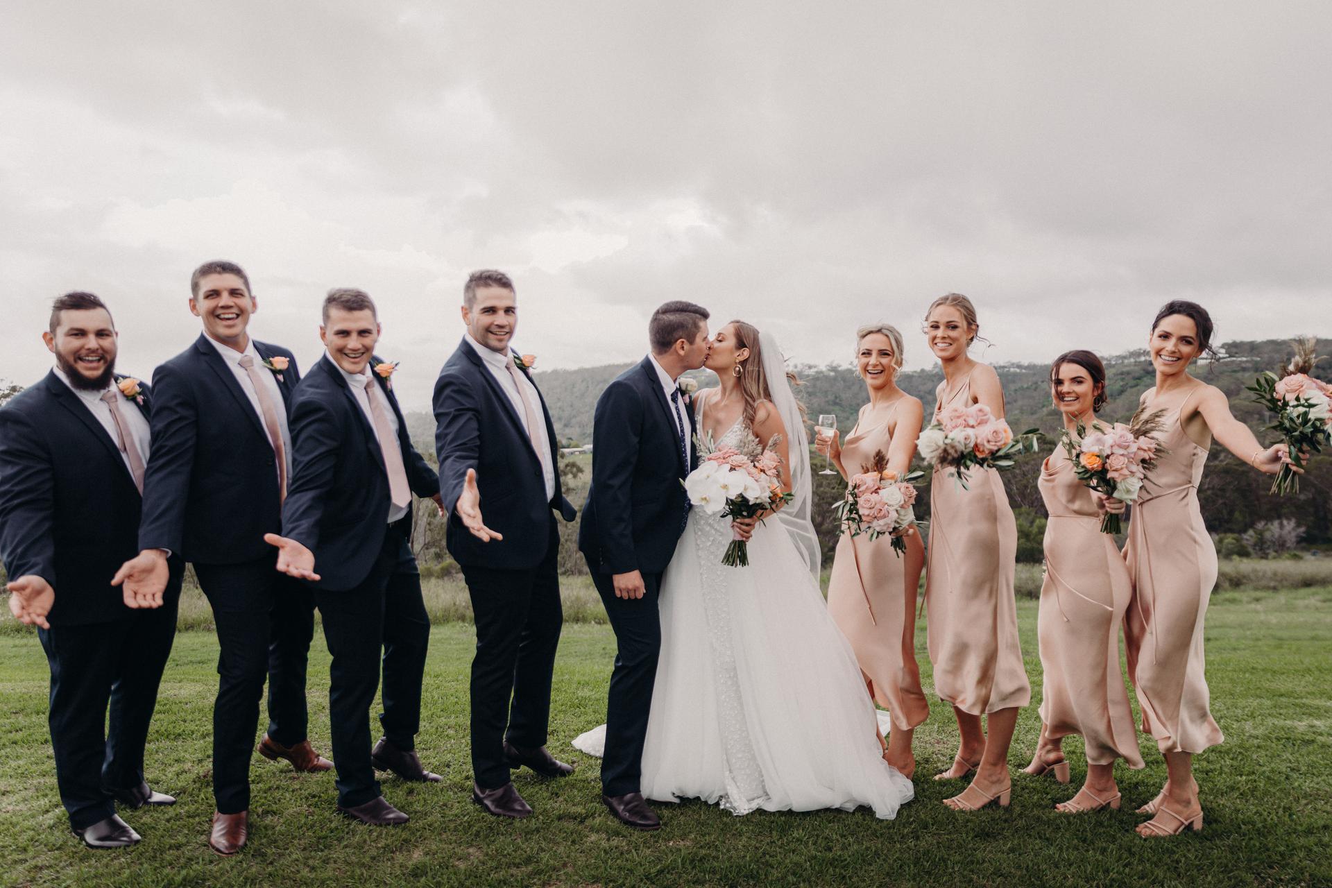 Chelsea the bride and Brent the groom kiss in the center while surrounded by five groomsmen in dark suits with peach boutonnieres on the left and five bridesmaids in matching light peach dresses holding bouquets on the right at Preston Peak Winery Functions Centre outdoors on grass with hills in the background under a cloudy sky.