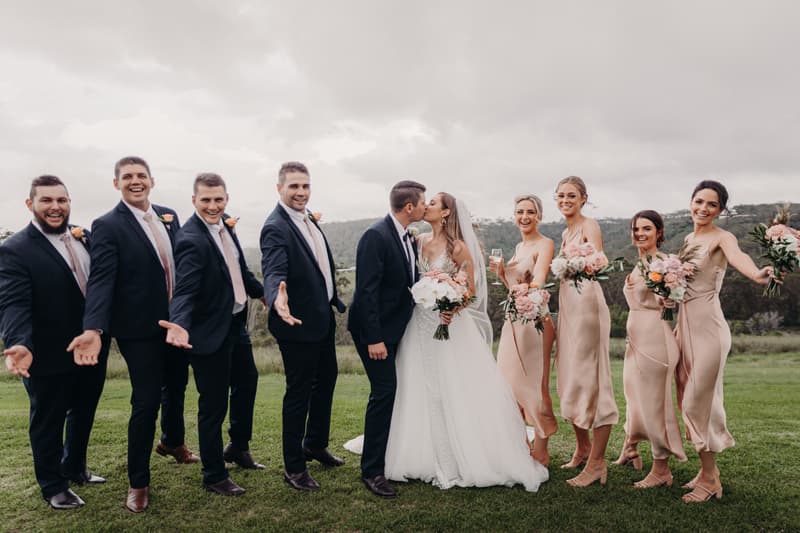 Chelsea the bride and Brent the groom kiss in the center while surrounded by five groomsmen in dark suits with peach boutonnieres on the left and five bridesmaids in matching light peach dresses holding bouquets on the right at Preston Peak Winery Functions Centre outdoors on grass with hills in the background under a cloudy sky.