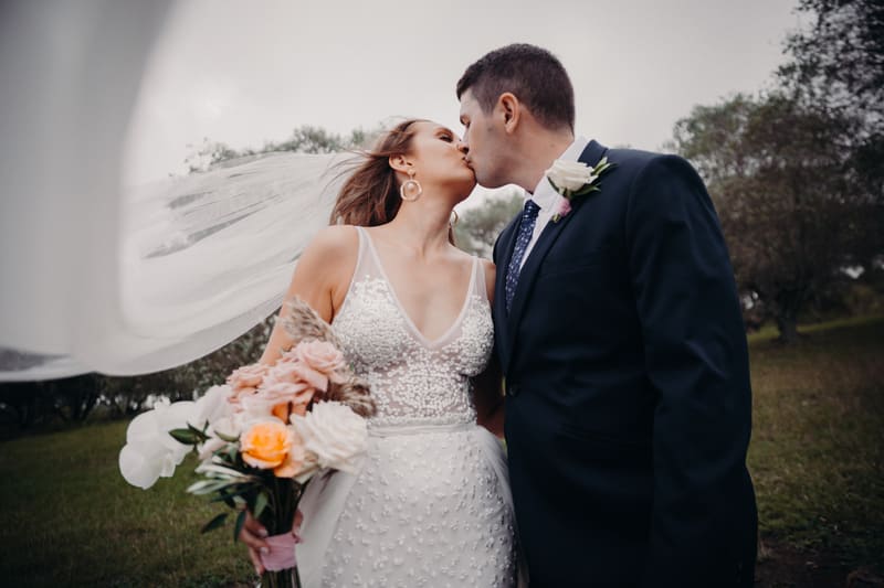 Chelsea the bride and Brent the groom share a kiss outdoors, with Chelsea holding a bouquet of flowers and her veil flowing behind her. Engagement party by She Said Yes.