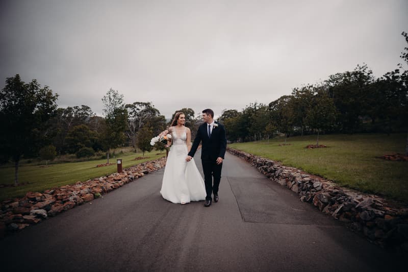 Chelsea in a white wedding gown holding a bouquet and Brent in a black suit holding hands while walking down a paved path bordered by stone walls and trees at Preston Peak Winery Functions Centre.