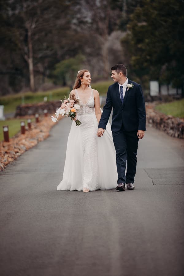 Chelsea the bride and Brent the groom walk hand in hand down a paved path at Preston Peak Winery Functions Centre. Chelsea wears a white wedding gown and holds a bouquet of flowers, while Brent is dressed in a dark suit with a boutonniere.