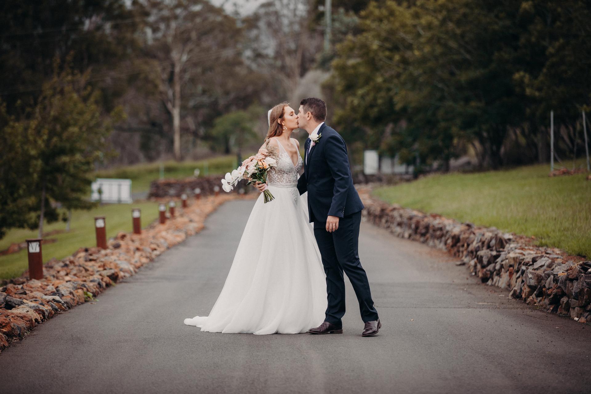 Chelsea the bride and Brent the groom kiss on a paved road lined with rocks and greenery at Preston Peak Winery Functions Centre during their couple portraits session.