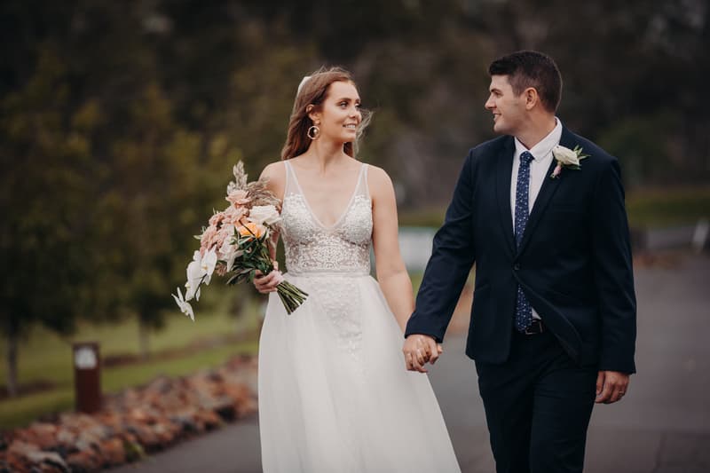 Chelsea the bride and Brent the groom walk hand in hand outdoors at Preston Peak Winery Functions Centre, with Chelsea holding a bouquet of flowers and both looking at each other.