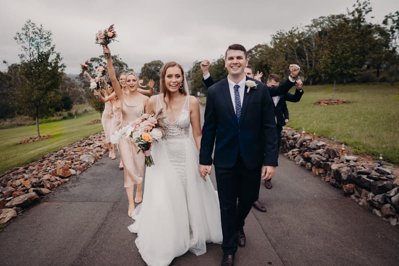 Chelsea and Brent walk hand in hand on a paved path at Preston Peak Winery Functions Centre, followed by bridesmaids in blush dresses holding bouquets and groomsmen in dark suits raising their fists in celebration.