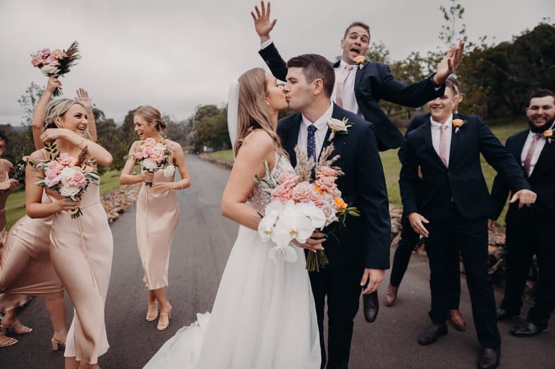 Chelsea the bride and Brent the groom kiss while surrounded by bridesmaids in blush dresses holding bouquets and groomsmen in dark suits with pink ties at Preston Peak Winery Functions Centre.