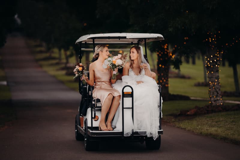 The bride Chelsea in a white wedding gown and a bridesmaid in a light pink dress sit together on the back of a golf cart at Preston Peak Winery Functions Centre, each holding a bouquet and a glass of wine, with trees wrapped in fairy lights in the background.
