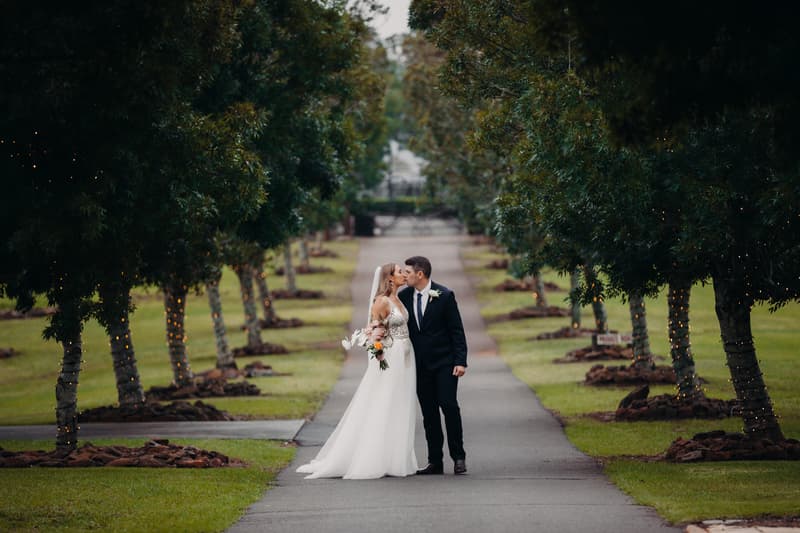 Chelsea and Brent kiss while standing on a tree-lined path at Preston Peak Winery Functions Centre, with trees wrapped in string lights on either side.