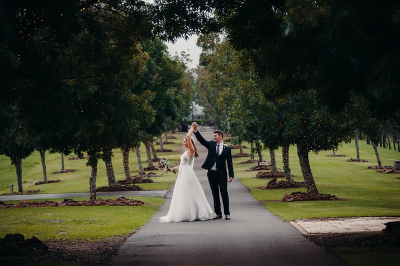 Chelsea and Brent stand on a paved path lined with trees wrapped in fairy lights at Preston Peak Winery Functions Centre, with Brent holding Chelsea's hand up as she twirls in her wedding dress holding a bouquet.