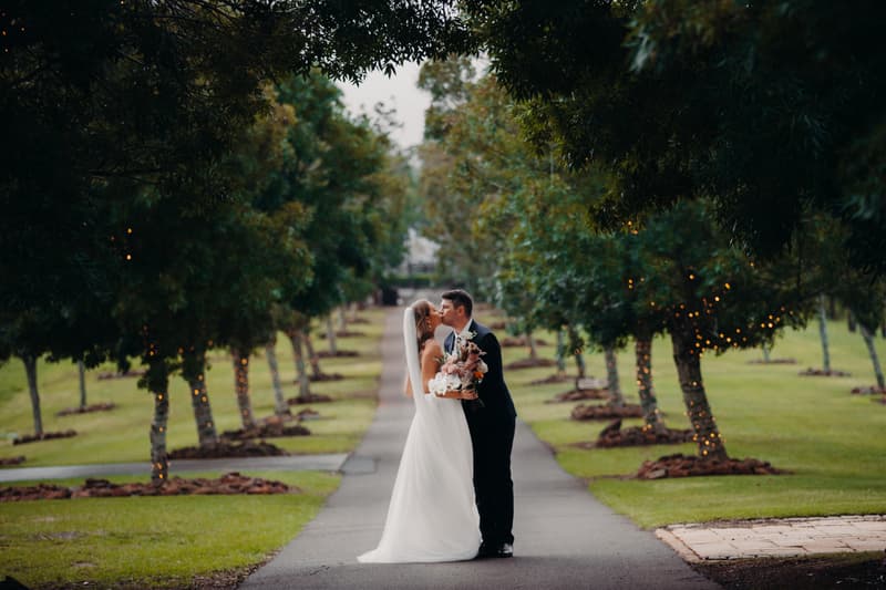 Chelsea and Brent share a kiss on a tree-lined path at Preston Peak Winery Functions Centre, with trees decorated with string lights on either side.