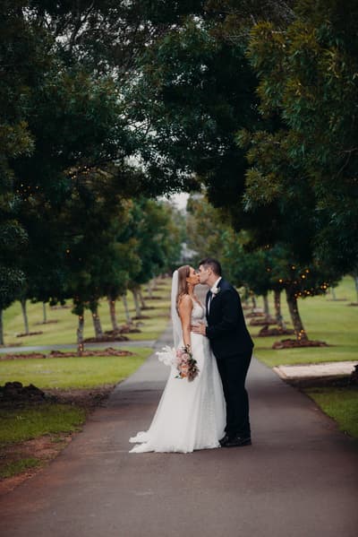 Chelsea and Brent kiss on a tree-lined path, with Chelsea holding a bouquet and wearing a wedding gown and veil, and Brent in a dark suit. ABIA 2018 finalist wedding photography by She Said Yes.