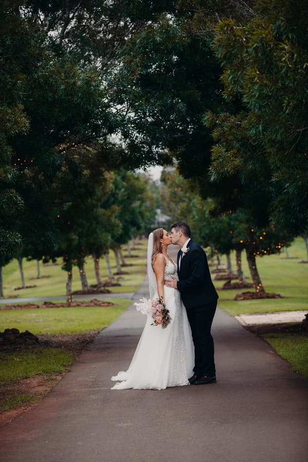 Chelsea and Brent kiss on a tree-lined path at Preston Peak Winery Functions Centre, with Chelsea holding a bouquet and wearing a wedding gown and veil, and Brent in a dark suit.