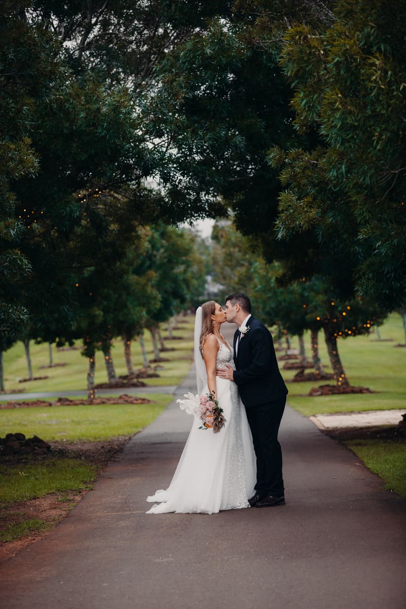 Chelsea and Brent kiss on a tree-lined path, with Chelsea holding a bouquet and wearing a wedding gown and veil, and Brent in a dark suit. Brisbane wedding photography by She Said Yes.