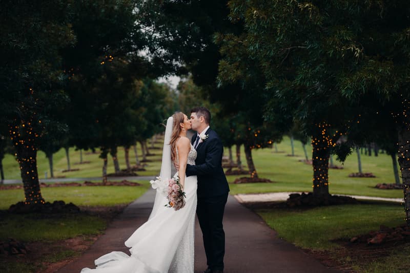 Chelsea the bride and Brent the groom kiss on a tree-lined path at Preston Peak Winery Functions Centre, with trees wrapped in string lights and green grass surrounding them.