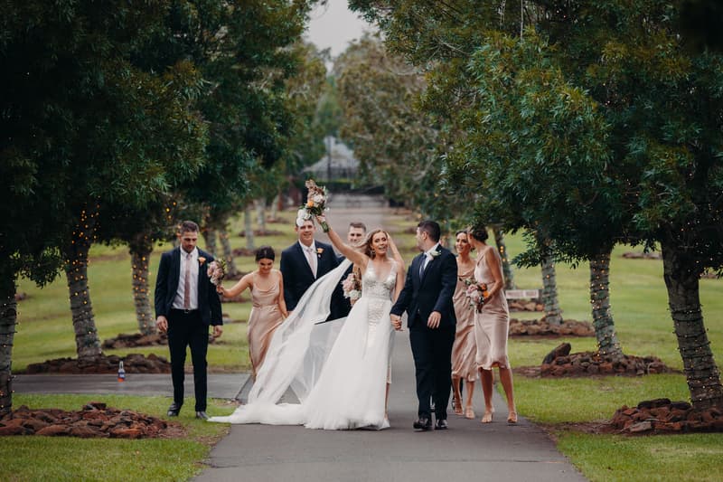 Chelsea the bride and Brent the groom walk hand in hand along a tree-lined path at Preston Peak Winery Functions Centre, accompanied by bridesmaids in champagne dresses and groomsmen in dark suits.