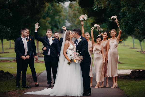 Chelsea the bride and Brent the groom kiss in the center of a tree-lined path, surrounded by bridesmaids in blush dresses holding bouquets and groomsmen in dark suits with peach boutonnieres. Brisbane videographer by She Said Yes.