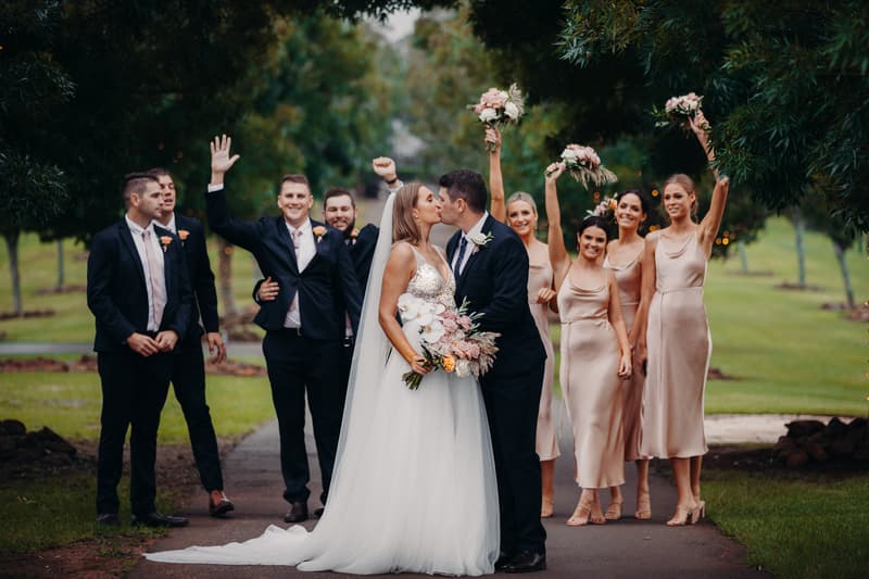 Chelsea the bride and Brent the groom kiss in the center of a tree-lined path at Preston Peak Winery Functions Centre, surrounded by bridesmaids in blush dresses holding bouquets and groomsmen in dark suits with peach boutonnieres.