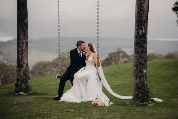 Bride Chelsea and groom Brent sit on a swing between two trees at Preston Peak Winery Functions Centre, sharing a kiss. Chelsea wears a white wedding dress with a long veil, and Brent is in a dark suit. A bouquet lies on the grass in front of them.