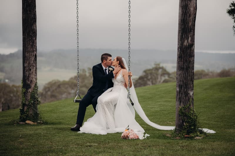 Bride Chelsea and groom Brent sit on a swing between two trees at Preston Peak Winery Functions Centre, sharing a kiss. Chelsea wears a white wedding dress with a long veil, and Brent is in a dark suit. A bouquet lies on the grass in front of them.