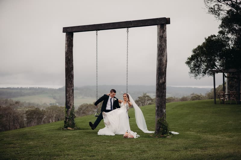 Bride Chelsea and groom Brent sit together on a wooden swing at Preston Peak Winery Functions Centre with a scenic grassy landscape and distant hills in the background. Chelsea wears a white wedding gown and veil, and Brent wears a dark suit with a boutonniere.