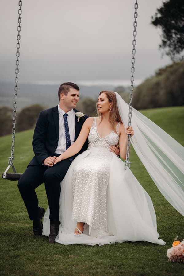 Chelsea and Brent sit together on a swing at Preston Peak Winery Functions Centre, with Chelsea wearing a white wedding dress and veil and Brent in a dark suit with a boutonniere.