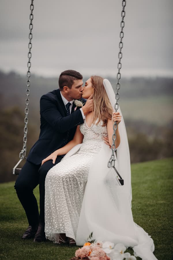Chelsea and Brent kiss while sitting on a swing at Preston Peak Winery Functions Centre, with Chelsea wearing a white wedding dress and veil and Brent in a dark suit.