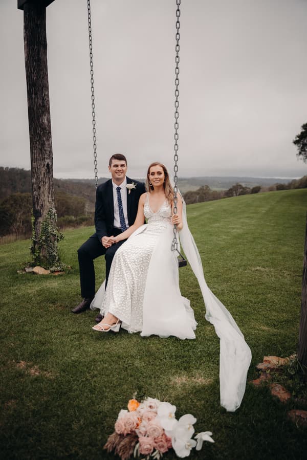 Chelsea and Brent sit together on a wooden swing at Preston Peak Winery Functions Centre, with a scenic grassy landscape and hills in the background. Chelsea wears a white wedding dress with a long veil, and Brent wears a dark suit with a boutonniere. A bouquet of flowers lies on the grass in front of them.