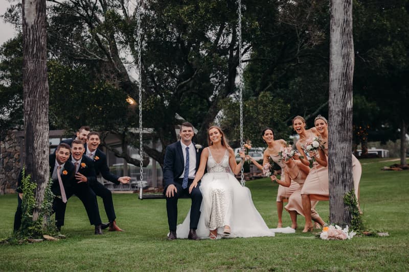 Chelsea and Brent sit together on a swing at Preston Peak Winery Functions Centre, surrounded by bridesmaids in blush dresses holding bouquets on the right and groomsmen in dark suits with orange boutonnieres on the left, all posing playfully on the grass outdoors.