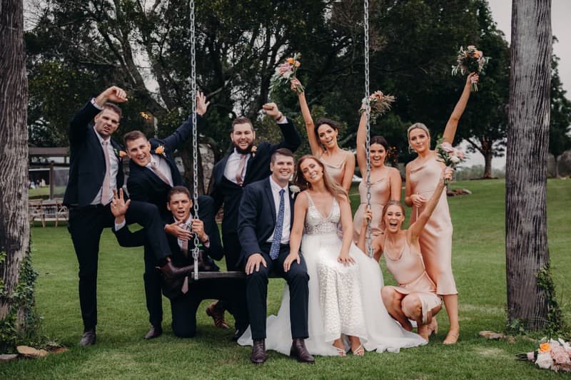 Chelsea the bride and Brent the groom sit on a swing at Preston Peak Winery Functions Centre surrounded by bridesmaids in matching blush dresses holding bouquets and groomsmen in dark suits with blush ties, all posing cheerfully on a grassy lawn with trees in the background.
