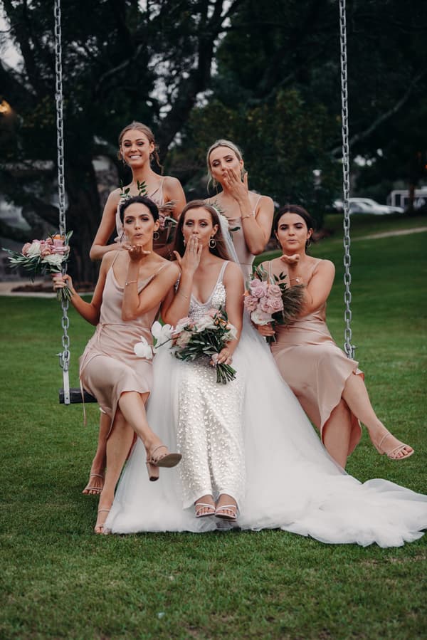 The bride Chelsea in a white wedding dress sits on a swing at Preston Peak Winery Functions Centre with four bridesmaids in matching beige dresses, all holding bouquets and blowing kisses towards the camera.