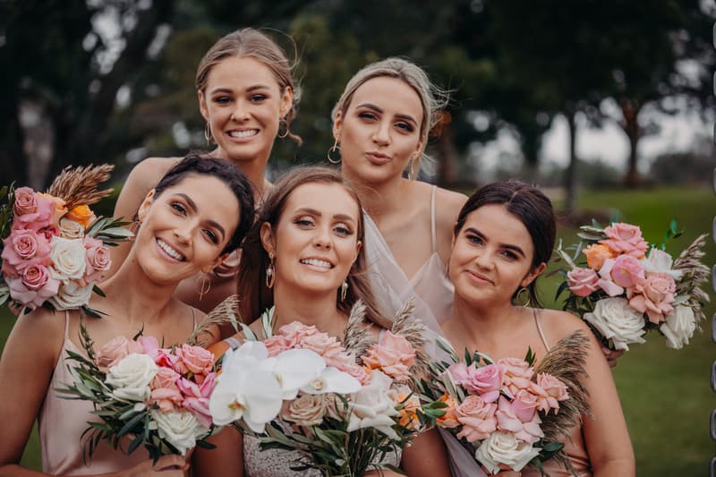 Five bridesmaids pose together outdoors at Preston Peak Winery Functions Centre, each holding bouquets of pink, white, and peach flowers with greenery.