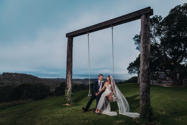Chelsea and Brent sit together on a large wooden swing at Preston Peak Winery Functions Centre, with a scenic hillside and trees in the background. Chelsea wears a white wedding gown and veil, holding a bouquet, while Brent wears a dark suit.