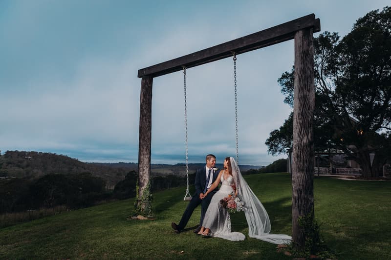 Chelsea and Brent sit together on a large wooden swing at Preston Peak Winery Functions Centre, with a scenic hillside and trees in the background. Chelsea wears a white wedding gown and veil, holding a bouquet, while Brent wears a dark suit.
