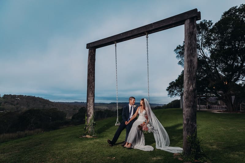 Chelsea and Brent sit together on a large wooden swing, with a scenic hillside and trees in the background. Chelsea wears a white wedding gown and veil, holding a bouquet, while Brent wears a dark suit. Cinematic videography wedding by She Said Yes.