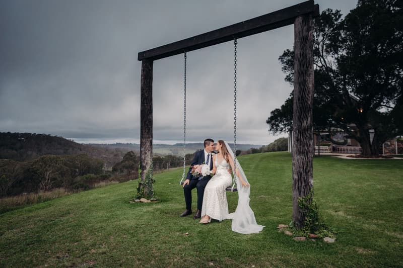 Chelsea and Brent sit together on a large wooden swing at Preston Peak Winery Functions Centre, with a scenic grassy hill and distant trees under a cloudy sky.